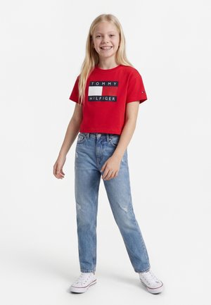 Χαμογελαστή κοπέλα με μακριά ξανθά μαλλιά, φοράει κόκκινο crop top Tommy Hilfiger, ανοιχτόχρωμα μπλε τζιν και λευκά αθλητικά παπούτσια, στέκεται μπροστά σε λευκό φόντο.