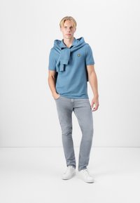 Lyseblå polo med en gul logo på brystet, kombinert med lysegrå jeans og hvite joggesko. En hettegenser er slengt over skuldrene.
