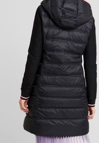 Zwarte puffer vest met capuchon, voorzien van een doorgestikt ontwerp, elastische manchetten met rode en witte strepen, en een lange, rechte snit.