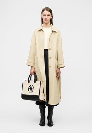 Woman wearing beige long coat, black skirt, and black boots, holding beige and black tote bag with AB logo on white background.