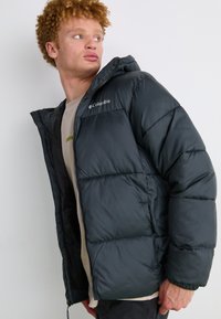 Schwarze Steppjacke mit Kapuze, mit einem ge quilteten Design, Reißverschluss und Columbia-Logo auf der linken Brust. Weicher, leichter Stoff.