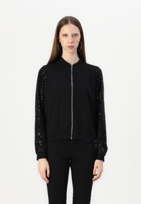 Schwarze Bomberjacke mit Reißverschluss vorne, aus mattem Stoff und langen Ärmeln mit Pailletten. Gerippte Bündchen und Saum. Insgesamt glatte Textur.