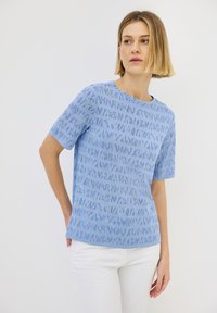 Modrá krátká tričková blůza s texturovaným vzorem abstraktních tvarů, kulatým výstřihem a volným střihem, kombinovaná s bílými kalhotami.