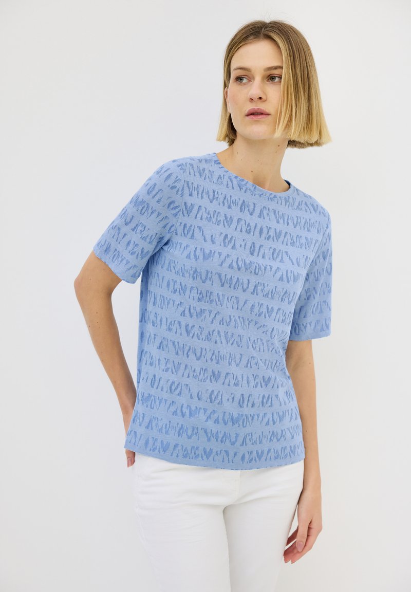 Modrá krátká tričková blůza s texturovaným vzorem abstraktních tvarů, kulatým výstřihem a volným střihem, kombinovaná s bílými kalhotami.