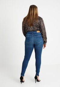 Mujer con cabello lacio castaño, vestida con una blusa de estampado de leopardo, jeans skinny azules y tacones altos negros, de pie contra un fondo blanco.