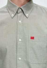 Gros plan d'une chemise gris clair à boutons blancs avec une poche poitrine comportant une petite étiquette rouge.