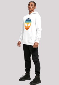 White hoodie with a vintage alien logo in blue, orange, and yellow stripes. Paired with black cargo pants and black sneakers.