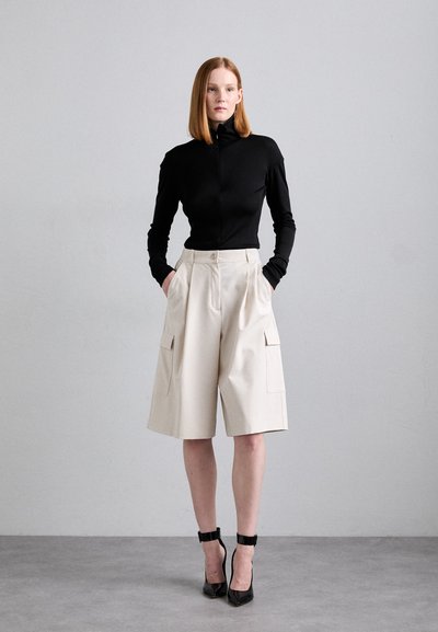 Musta pitkähihaiset paita korkealla kauluksella, yhdistettynä beigen värisiin pleated cargo-shortsihousuihin, joissa on sivutaskut. Mallilla on päällään mustat nilkkaketjulliset korot.
