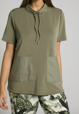 Top verde oliva de manga corta con un cuello alto, detalle de cordón y dos bolsillos frontales. Textura suave, ajuste casual, combinado con pantalones con estampado.