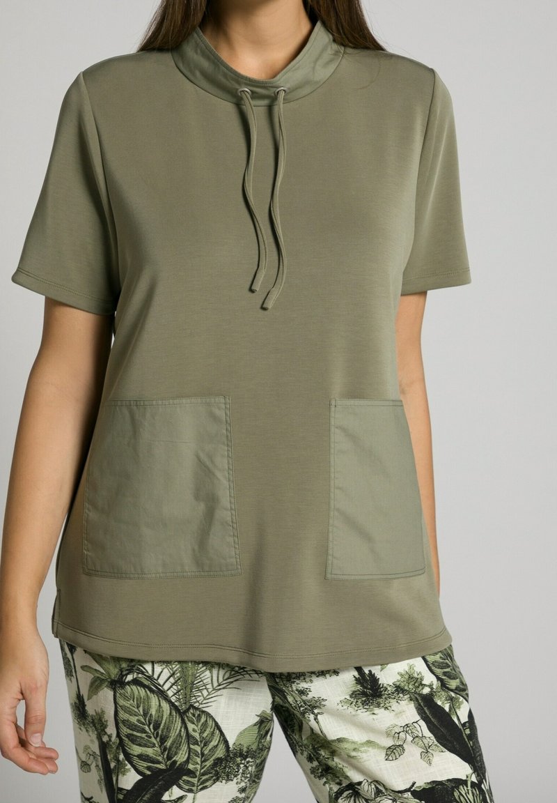 Top corto a maniche lunghe verde oliva con colletto alto, dettaglio con cordoncino e due tasche frontali. Texture liscia, vestibilità casual, abbinato a pantaloni a fantasia.