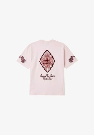 Camiseta rosa claro con diseños paisley burdeos en las mangas y un patrón central ornamentado en forma de diamante, además del texto "Change The Game Project X Paris".