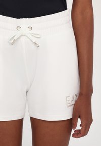 Pantalones cortos de algodón blanco con una cintura ajustable, que presentan un logo bordado en dorado en el lado inferior, textura suave y un corte relajado.