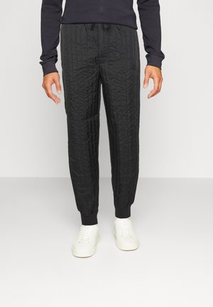 Mand iført sorte quiltede joggerbukser, sort langærmet top og hvide sneakers, stående foran en ensfarvet hvid baggrund.