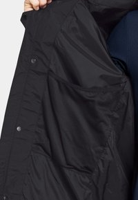 Sort jakke indvendig med glat, letvægtsstof og en mesh-lomme. Har trykknapper og et robust design for funktionalitet og komfort.
