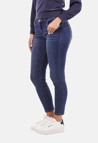 Jeans skinny blu con una texture in denim liscia, dotati di vita media, tasche frontali e chiusura con bottone, abbinati a sneaker bianche.