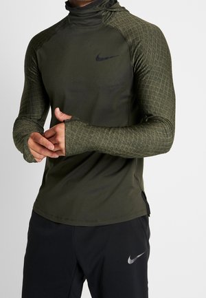 Homme portant un haut de sport texturé à manches longues vert foncé avec des passe-pouces et un pantalon noir Nike, ajustant sa main gauche.