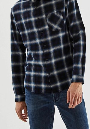 Uomo che indossa una camicia a quadri nera e blu a maniche lunghe con tasca frontale abbinata a jeans blu, in piedi davanti a uno sfondo semplice.