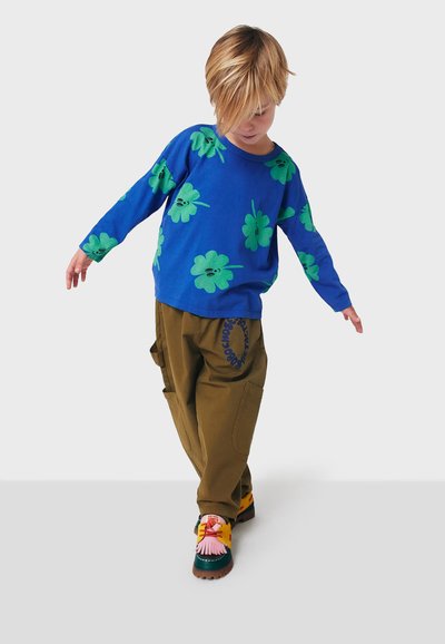 Blauses langärmliges Hemd mit grünen Blumenprints, lockere braune Cargo-Hosen mit Seitentaschen und bunte Schuhe mit unterschiedlichen Texturen und Mustern.