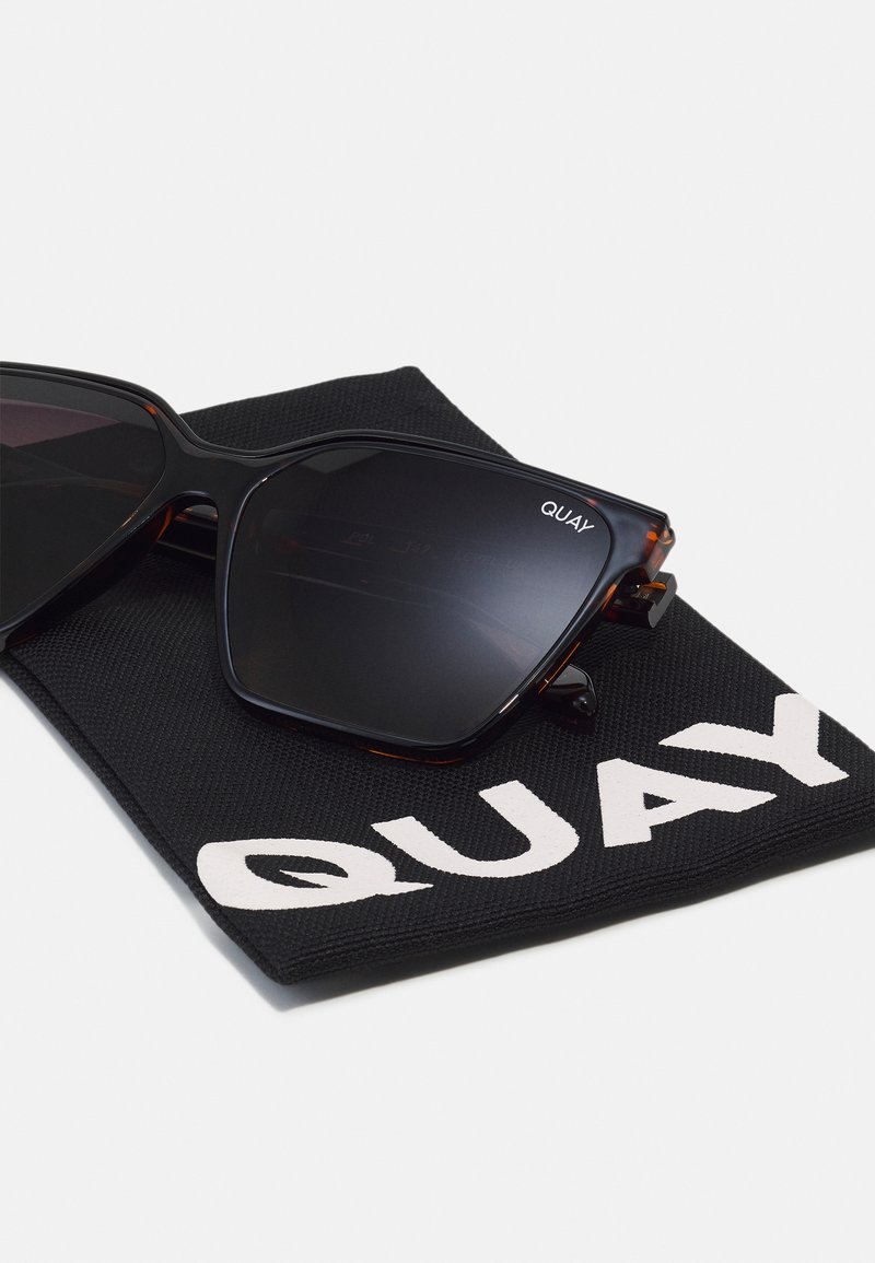 Black sunglasses with dark lenses, angular frame design featuring brown tortoiseshell accents, resting on a black cloth pouch with "QUAY" branding.