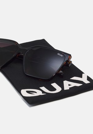 Zwarte cat-eye zonnebril met donkere glazen die rust op een zwarte stoffen etui bedrukt met de witte merknaam Quay.