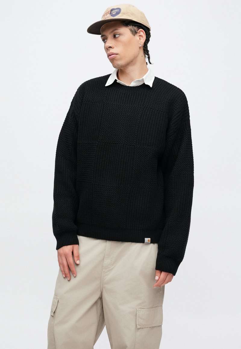 Sort strikket sweater med et tekstureret gittermønster; afslappet pasform, rund hals, ribbet manchetter og kant. Bæres med beige cargobukser og en kasket.