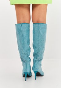 Botine turqoise până la genunchi, cu o suprafață texturată și cu ținte argintii, vârf ascuțit și toc stiletto. Botinele au un design ajustat și detalii cu cusături.