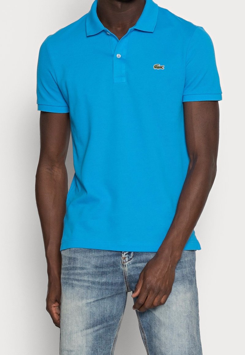 Homme portant un polo bleu vif à manches courtes avec un petit logo crocodile sur la poitrine et un jean bleu délavé.