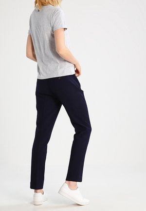 Personne portant un t-shirt gris à manches courtes, un pantalon bleu marine et des baskets blanches, debout de profil sur un fond blanc uni.