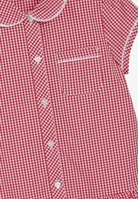 Punainen ja valkoinen ruudullinen puuvillapaida, jossa on pyöreä kaulus, lyhyet hihat ja tasku. Varustettu valkoisilla nappikiinnityksillä ja reunatunneilla.