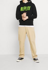 Sudadera negra con gran texto verde "REPLAY", pantalones beige y zapatillas negras con punteras blancas. La sudadera tiene cordones y acentos laterales.