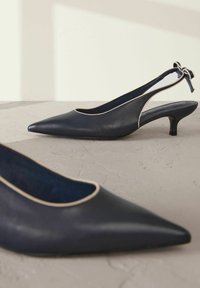 Tummansininen nahkainen slingback-korkokenkä, jossa on teräväkärkinen muotoilu, kerma värinen reuna ja rusetti koristeena kantapäällä. Strukturisoitu pinta. Alhaiset kitten-korkokengät.