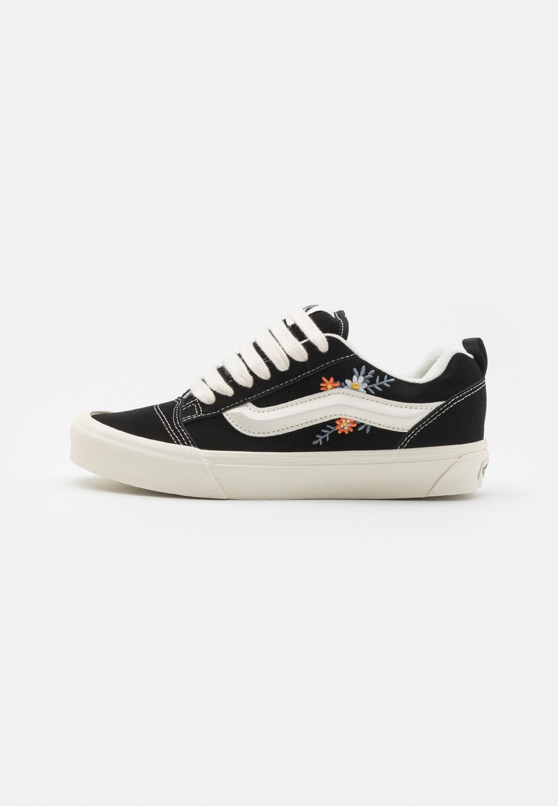 Svart canvas sneakers med hvite lærdetaljer, kremfargede snøringer og blomsterbroderi i blå, oransje og gult på utsiden.