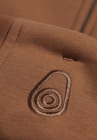 Tejido marrón con una textura acanalada que presenta un logo de espiral bordado en un hilo marrón más oscuro, situado cerca de la zona del zip.