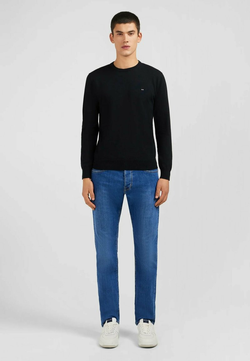 Pull en tricot noir à manches longues, col rond et petit logo sur la poitrine, associé à un jean bleu clair et des baskets blanches.