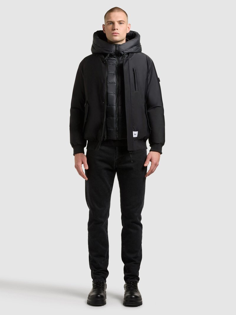 Czarna ocieplana kurtka z wysokim kołnierzem i dużym kapturem, noszona na puchowej kamizelce. Do tego dopasowane czarne jeansy i masywne czarne buty.