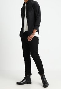 Camicia in denim nero sopra una t-shirt bianca, pantaloni slim-fit neri e stivali chelsea neri lucidi. Tra le caratteristiche ci sono tasche con bottoni e maniche arrotolate.