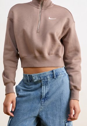 Osoba ubrana w skróconą brązową bluzę Nike z zamkiem błyskawicznym z przodu i niebieskie dżinsy z wysokim stanem, ręce swobodnie przy bokach.