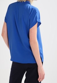 Blå kortärmad blus med uppvikta ärmar, slät textur, avslappnad passform och sidofickor, tillsammans med mörka byxor.