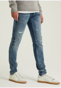 Persona vistiendo jeans ajustados de mezclilla azul desgastados y zapatillas blancas de caña baja con suelas de goma, de pie contra un fondo claro.