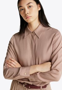 Jasnobrązowa koszula w paski z wywijanym kołnierzykiem, z przodem zapinanym na guziki i rękawami z mankietami. Gładka tekstura z akcentami złotych guzików.