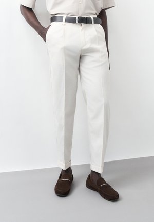 Mężczyzna w kremowych plisowanych spodniach, czarnym pasku i brązowych mokasynach stoi z rękami w kieszeniach na białym tle.