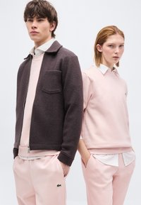 Νεαρός άνδρας και γυναίκα στέκονται δίπλα δίπλα, φορώντας ανοιχτό ροζ καθημερινά ρούχα· ο άνδρας φοράει σκούρο μπουφάν με φερμουάρ πάνω από ροζ μπλουζάκι.