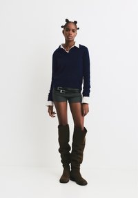 Pullover a maglia blu navy con colletto, indossato sopra una camicia bianca con colletto, abbinato a shorts grigi e stivali alti in pelle marrone.