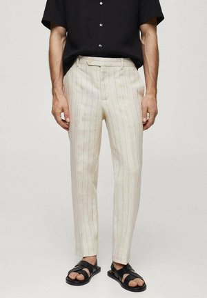 Hombre con pantalones de rayas verticales finas color crema, camisa negra de manga corta con botones y sandalias de cuero negras, de pie contra un fondo liso.