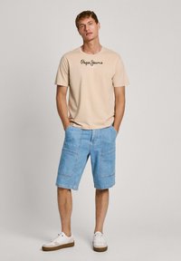 Camiseta beige con logo negro "Pepe Jeans London", combinada con pantalones cortos de mezclilla azul claros y zapatillas blancas con suelas marrones. Look limpio y casual.