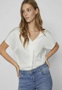 Blusa in satin bianco con scollatura a V e maniche corte, abbinata a jeans blu a vita alta. Texture morbida e vestibilità rilassata.