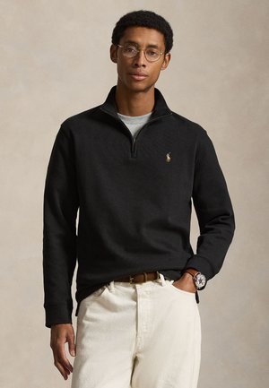 Mand iført runde briller, sort kvartlynlås-pullover med logo, hvide bukser, brun bælte og ur, står med den ene hånd i lommen.