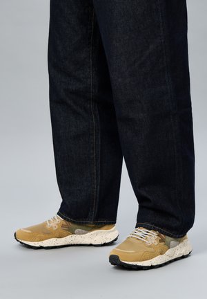 Piernas con vaqueros azul oscuro y zapatillas beige con suelas blancas salpicadas y cordones grises sobre un fondo gris claro.