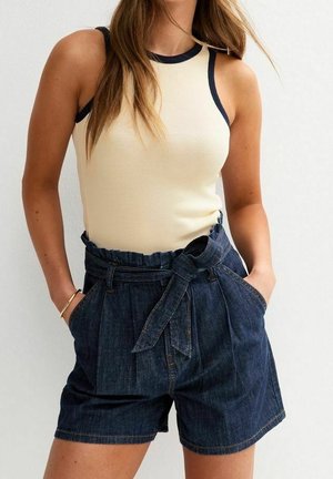 Kvinde iført ærmeløs cremefarvet top med sort kant og højtaljede mørke denimshorts med papirpose-talje og stofbælte, hænderne i lommerne.