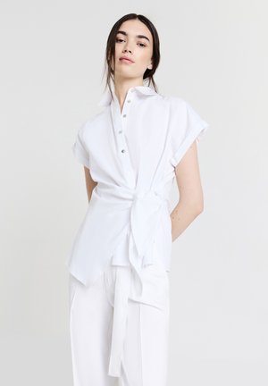 Chemise blanche à manches courtes avec un col, dotée de boutons et d'un détail noué à la taille. Le tissu semble lisse et léger.